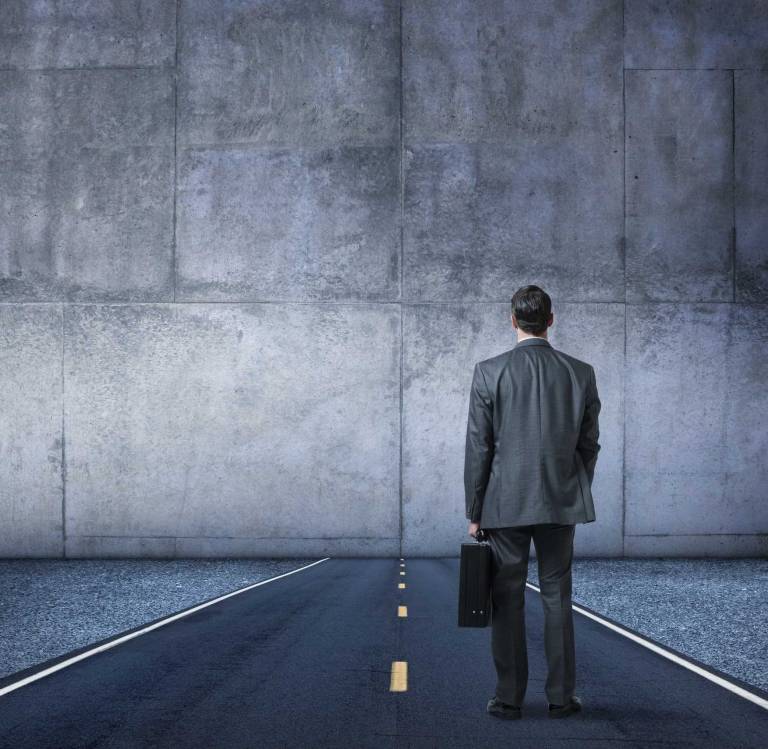 business professional holding a briefcase stands on a road facing a large concrete wall blocking the path forward, representing how changing buyer behavior and self-directed research create external challenges that stall traditional SaaS marketing and sales approaches
