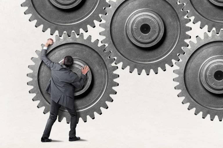 business professional pushes against oversized interlocking gears, symbolizing internal friction between marketing, sales, and product teams that prevents SaaS organizations from operating efficiently and achieving revenue alignment