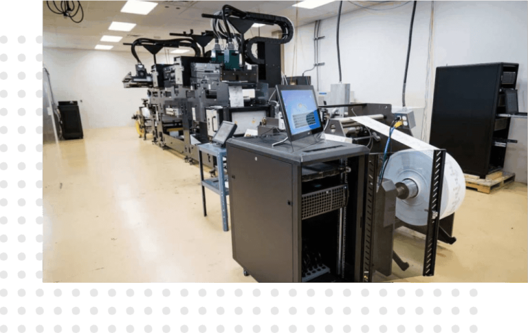 An industrial printing setup inside a well-lit room. The equipment includes large printing machines with various components and control panels.
